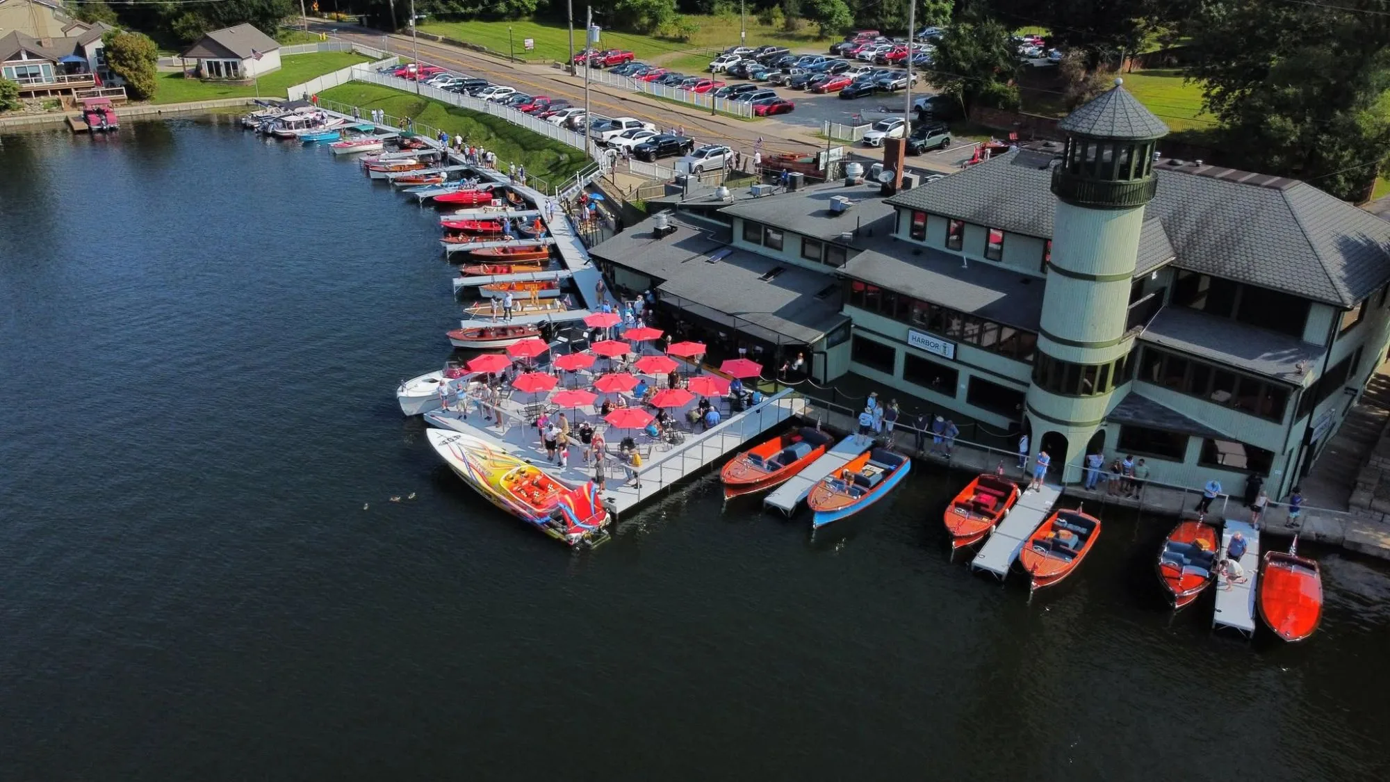 Aerial view of Harbor Lighthouse Grille on Portage Lakes: classic wooden boats lined up along the docks, red umbrellas on the floating patio, and the lighthouse tower rising over the green building