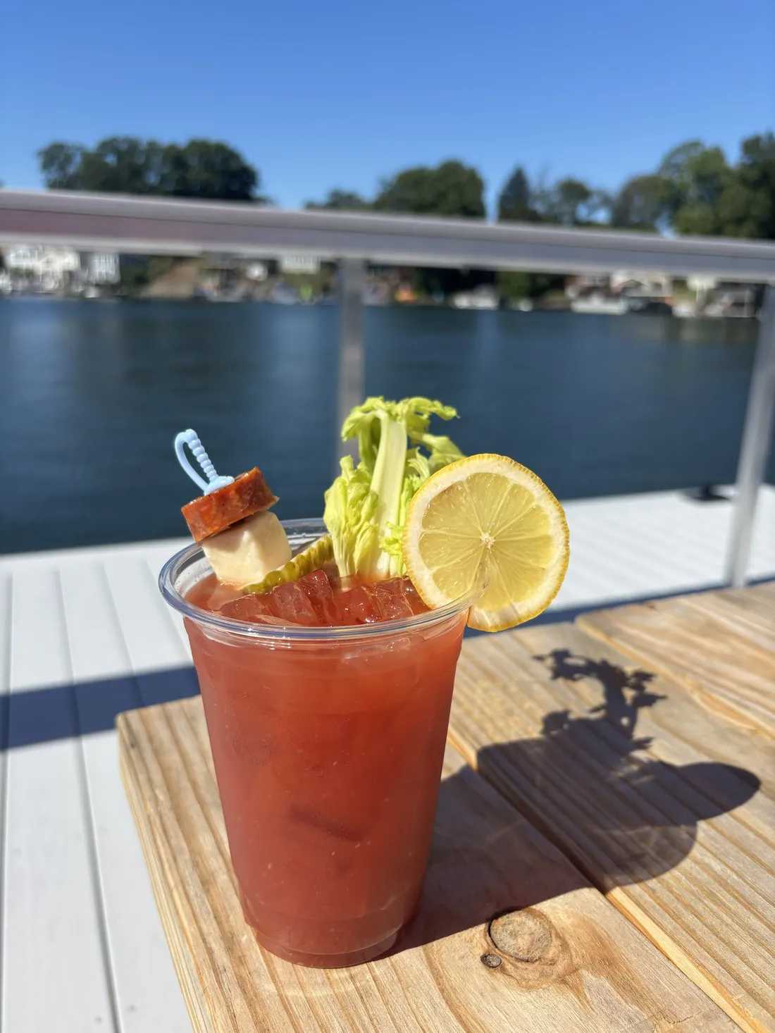 A Bloody Mary with a pepperoni and cheese skewer, celery and lemon, on a dock table over the lake